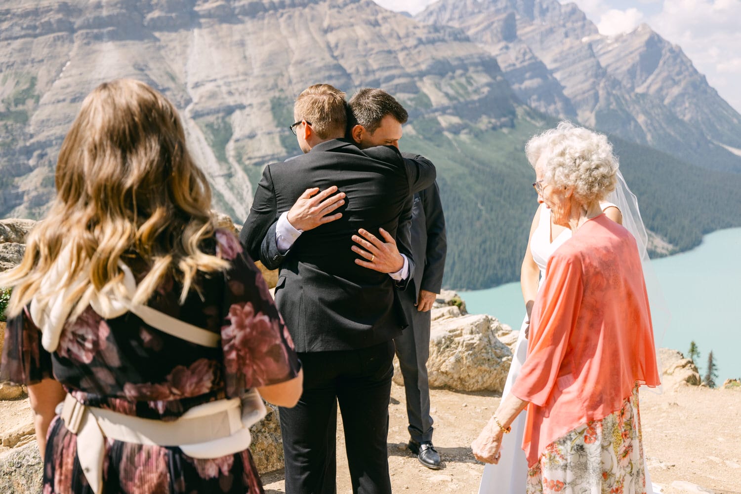 Two men embrace in a heartfelt moment during a scenic outdoor wedding at a cliffside overlooking a turquoise lake and mountains, surrounded by family and friends.