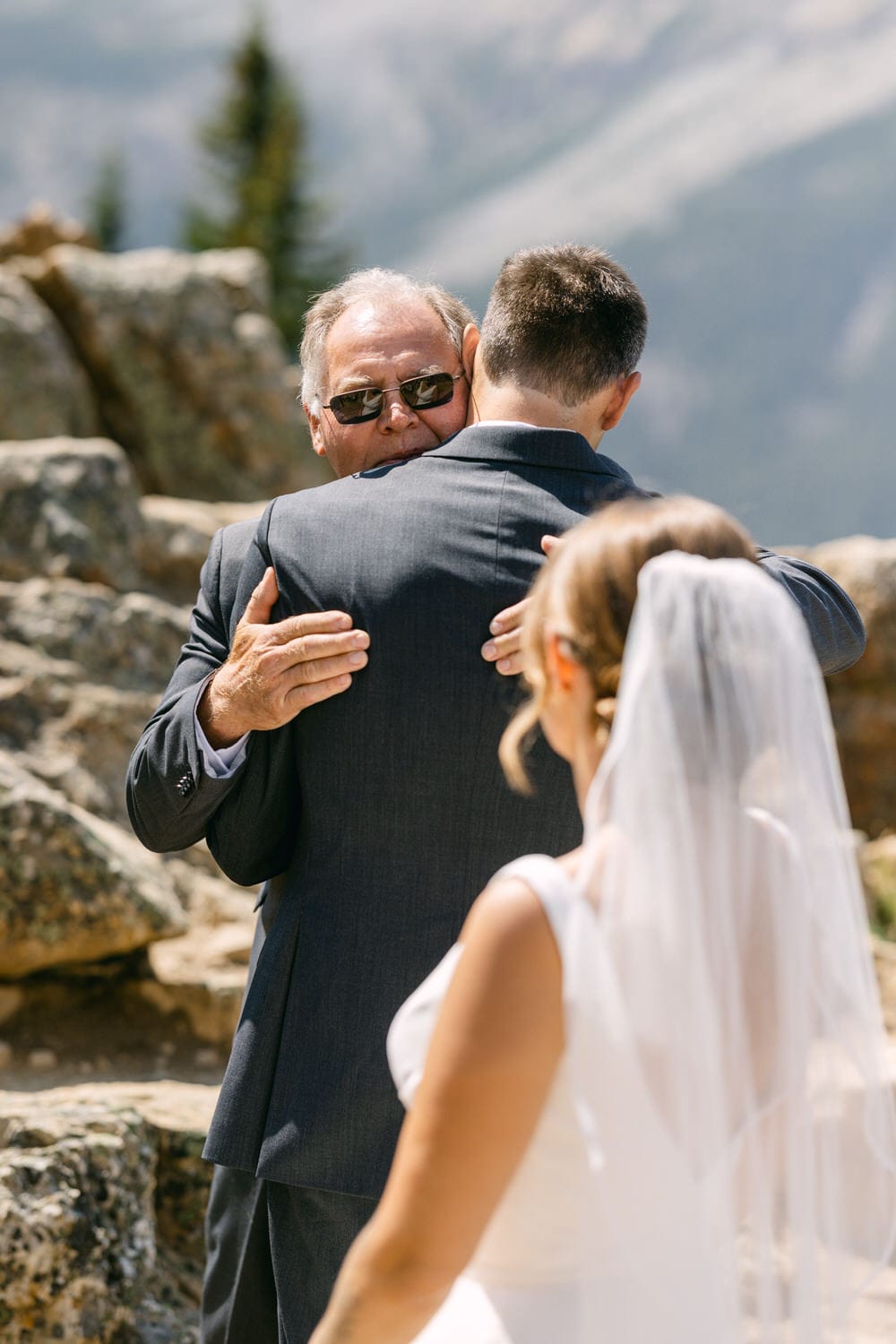 A formal-clad father hugs his son in a heartfelt moment during a wedding ceremony, with the bride visible in the background against a scenic mountainous backdrop.