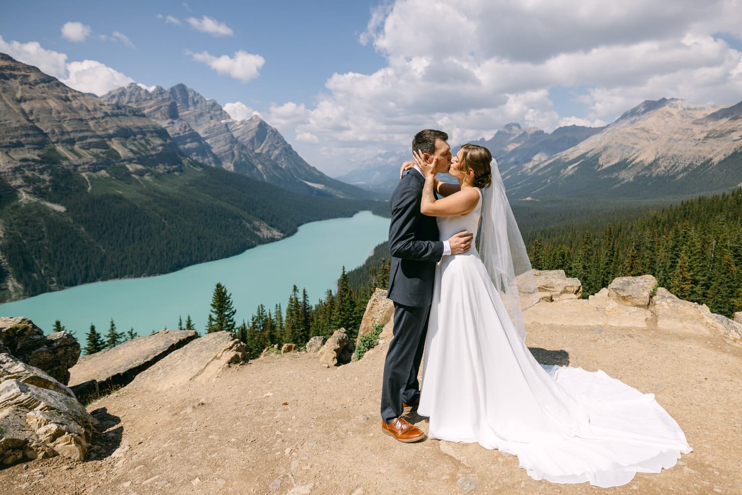 A couple shares a romantic kiss in a stunning mountainous landscape, with a turquoise lake in the background.