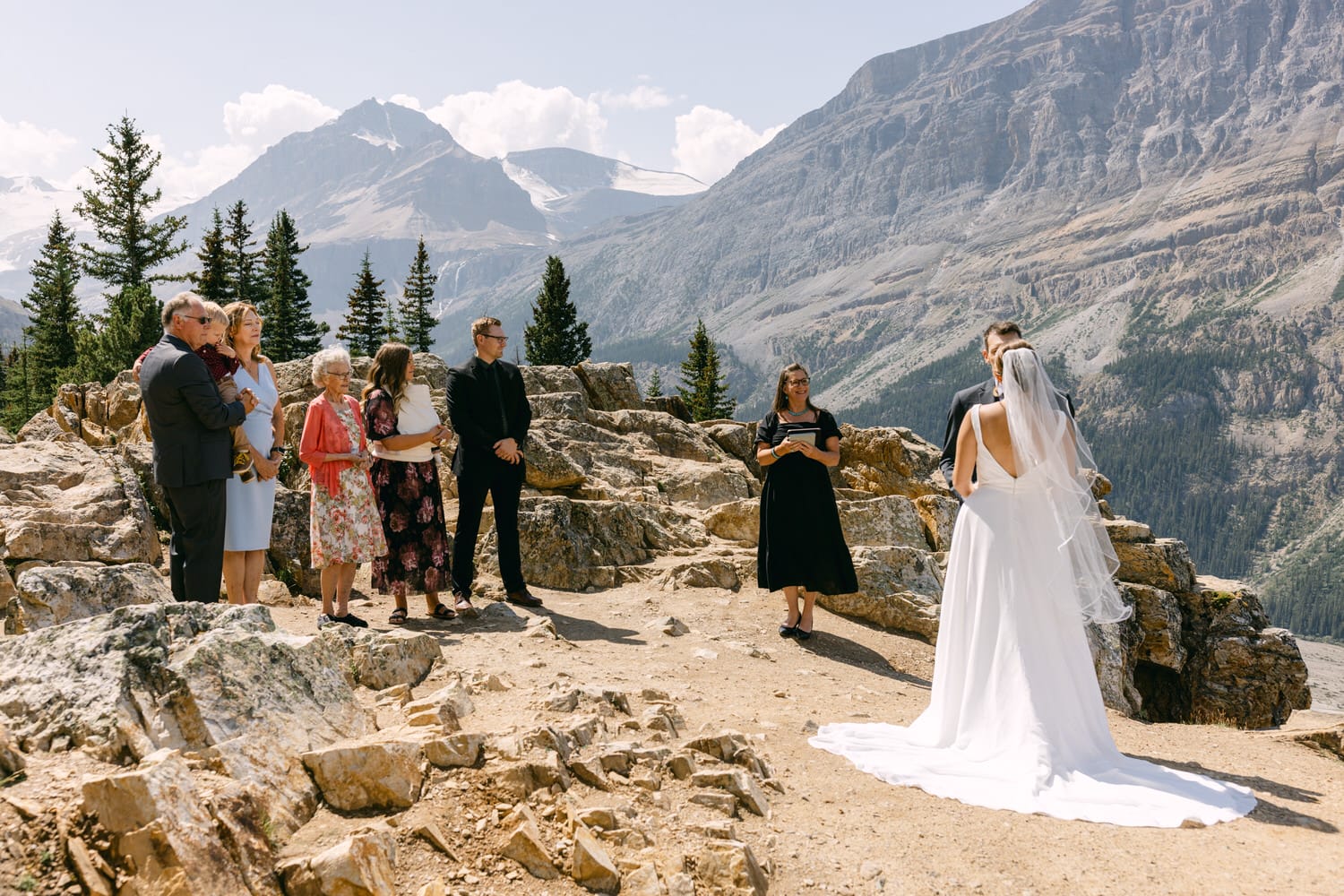 A bride and groom exchanging vows with a scenic mountain backdrop, surrounded by family and friends in a beautiful outdoor setting.