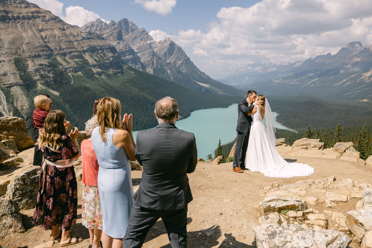 A couple sharing a kiss during their wedding ceremony at a scenic mountain viewpoint, surrounded by guests and a breathtaking landscape.