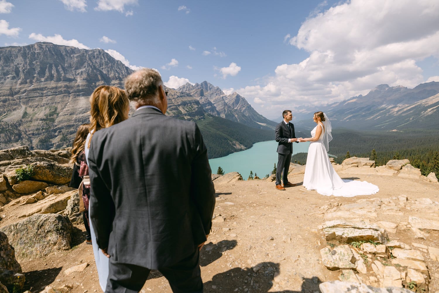 A couple exchanges vows on a rocky ledge overlooking a turquoise lake and majestic mountains, surrounded by guests.