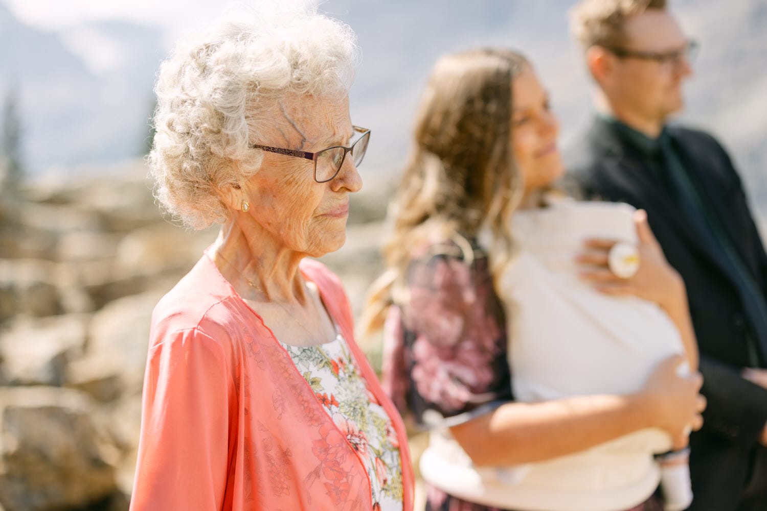 An elderly woman with curly hair and glasses, wearing a pink jacket, stands thoughtfully as a woman holding a baby and a man in a suit are seen in the background.