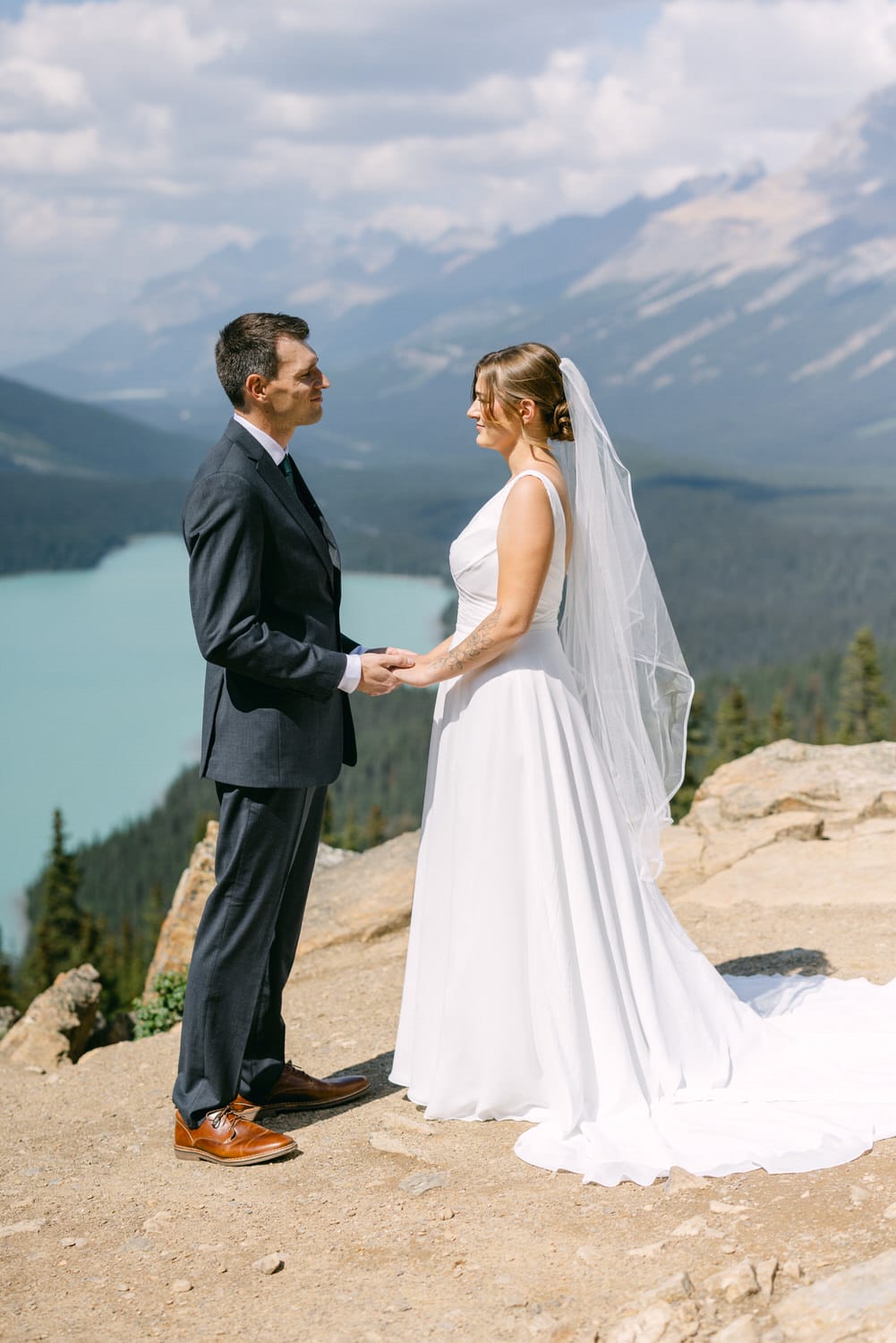 A couple exchanging vows on a scenic mountaintop with a lake and mountains in the background.