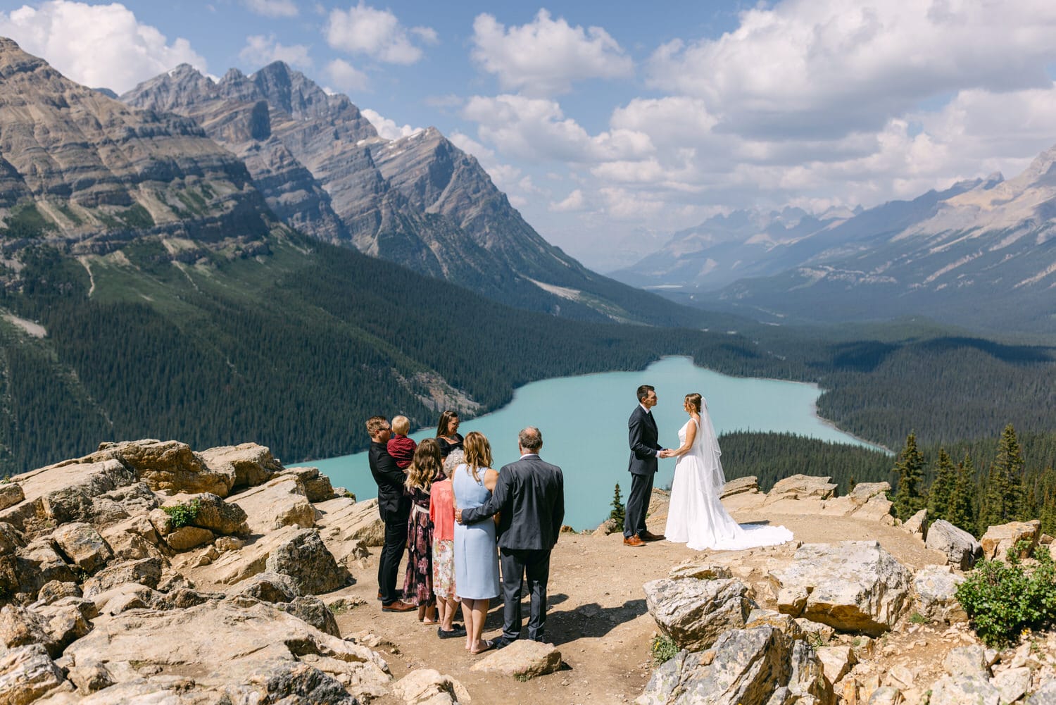 A couple exchanging vows atop a rocky cliff with breathtaking mountain and lake views, surrounded by family and friends.