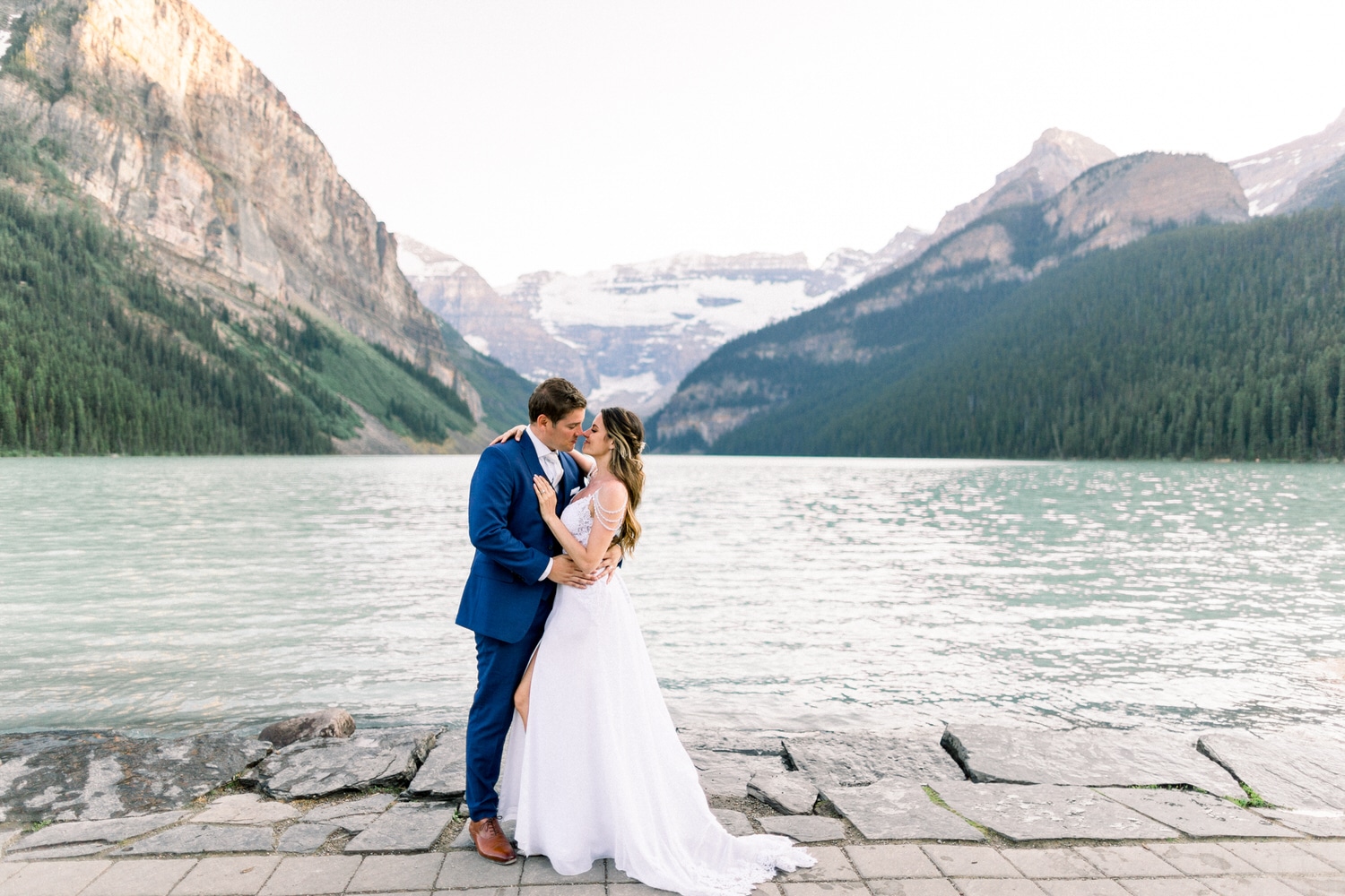 A couple embracing in wedding attire by a serene lake, surrounded by towering mountains and lush greenery.