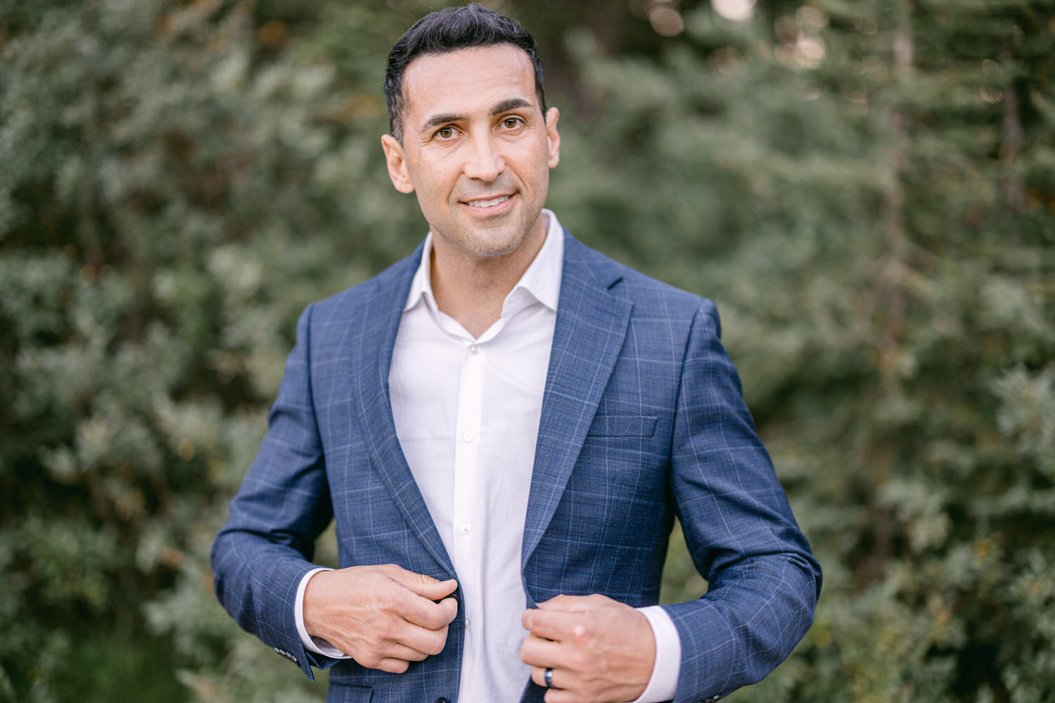 A well-dressed man adjusts his suit jacket while smiling in a natural setting with greenery in the background.