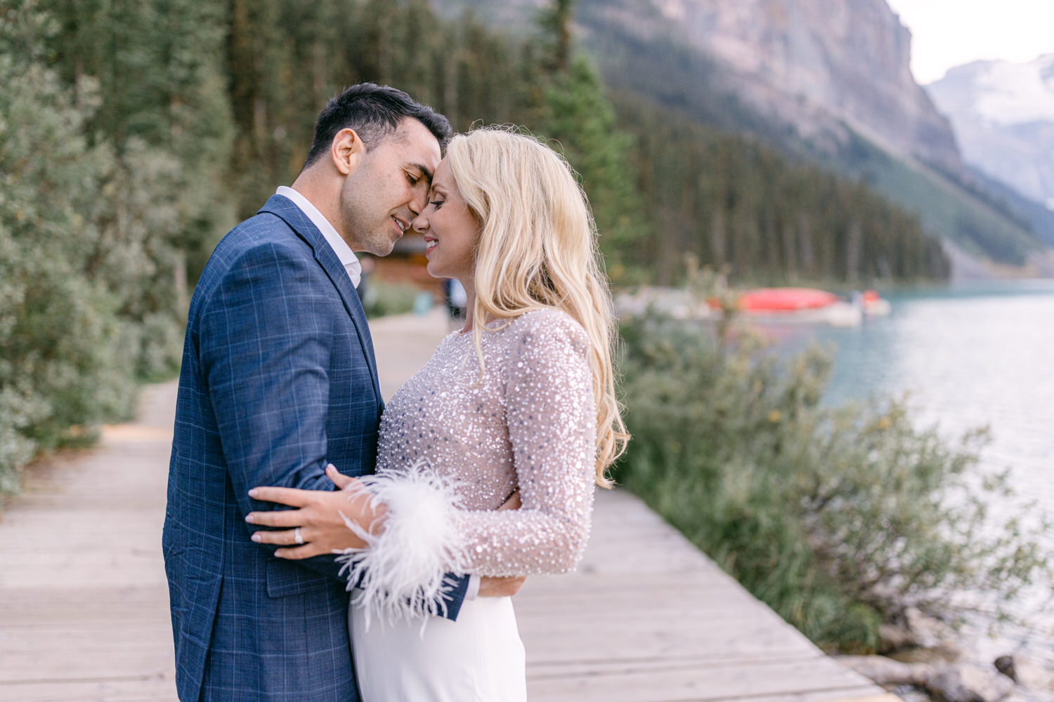 A couple shares an intimate moment on a scenic lakeside boardwalk, surrounded by lush trees and mountains.