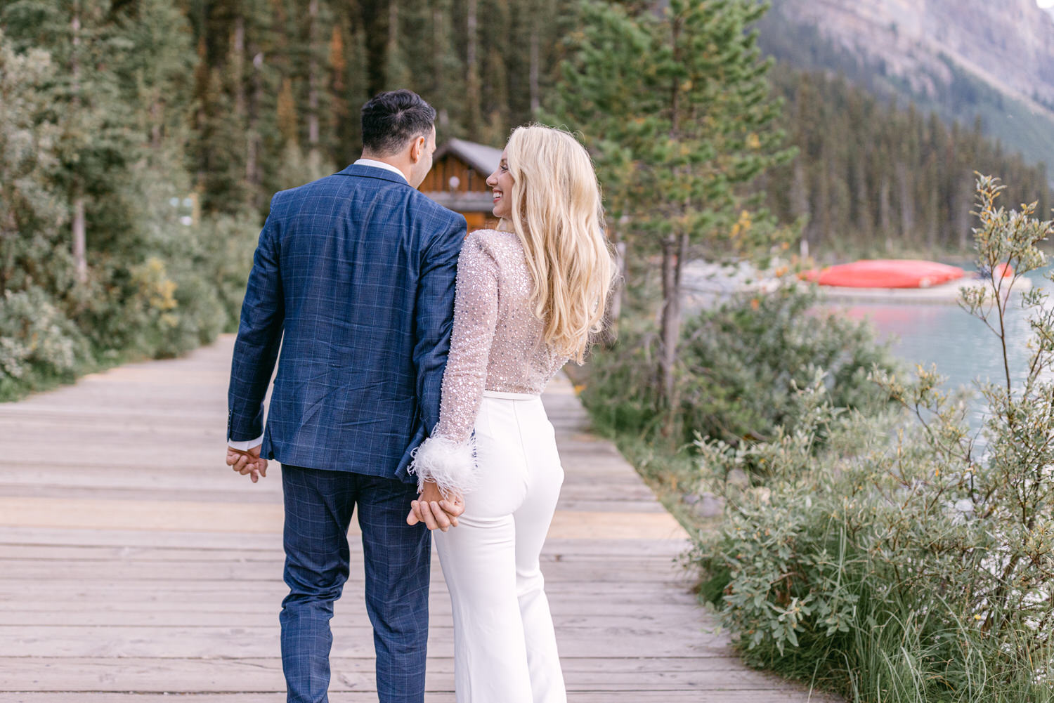 A couple walks hand in hand along a wooden pathway beside a serene lake, surrounded by trees and mountains, with the woman wearing a sparkling top and the man in a stylish suit.