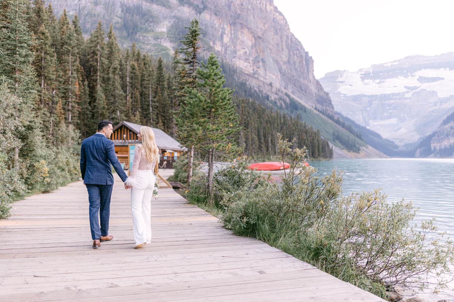 A couple dressed elegantly walks hand in hand along a wooden pathway beside a tranquil lake, surrounded by tall trees and mountains.