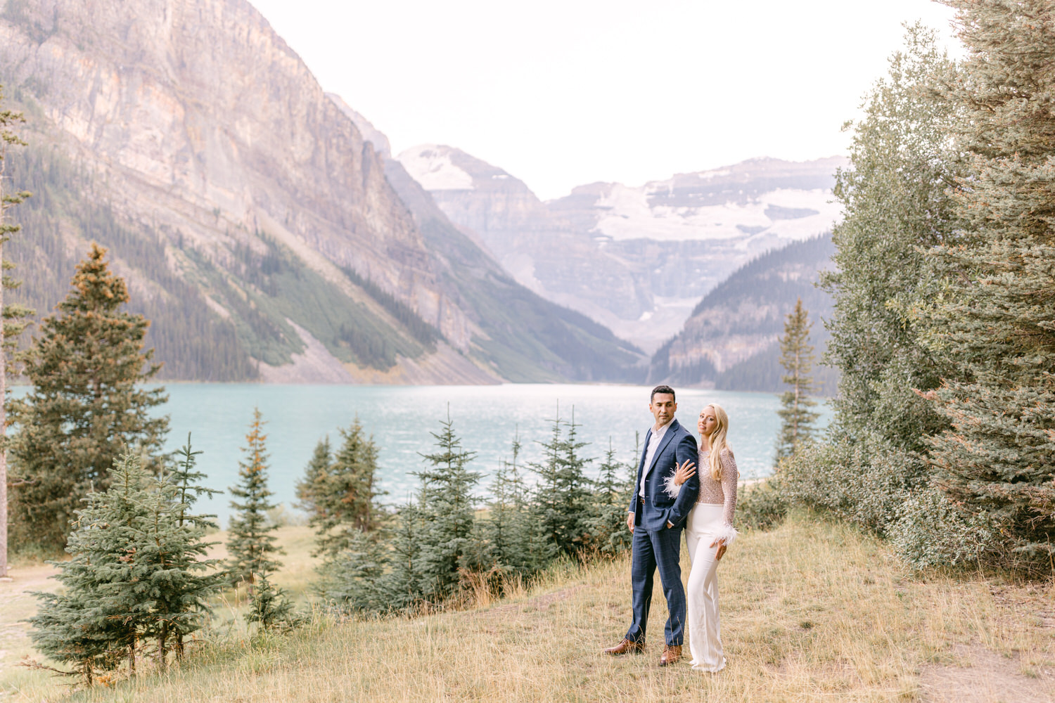 A couple stands together on the shore of Lake Louise, surrounded by lush greenery and majestic mountains in the background, evoking a romantic atmosphere.