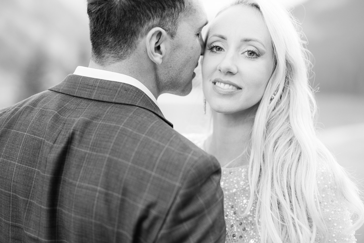 A close-up of a couple sharing a tender moment, with the man whispering to the woman who is smiling gently, set against a blurred natural background.