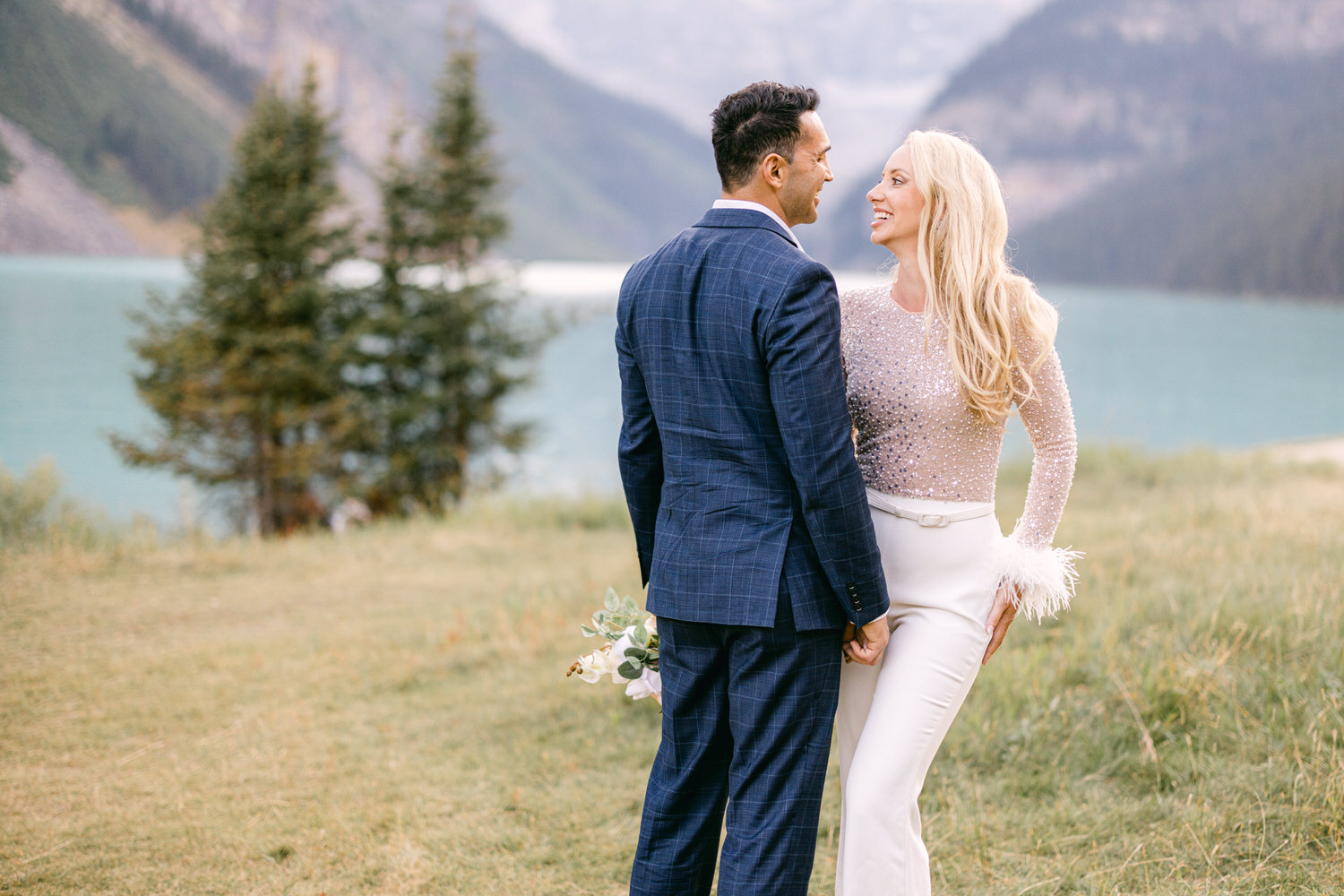 A happy couple in formal attire stands together by a serene lake, with mountains in the background, showcasing a romantic moment surrounded by nature.