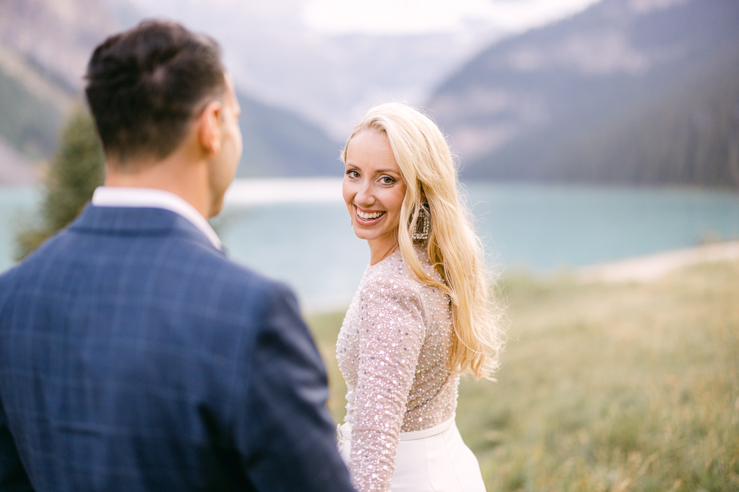 A smiling woman with long blonde hair and sparkling attire looks back at a man in a suit, set against a scenic lake and mountainous background.