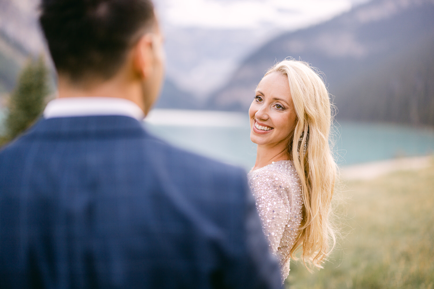 A woman in a sparkling dress smiles warmly at a man in a suit, set against a stunning lakeside backdrop with mountains.