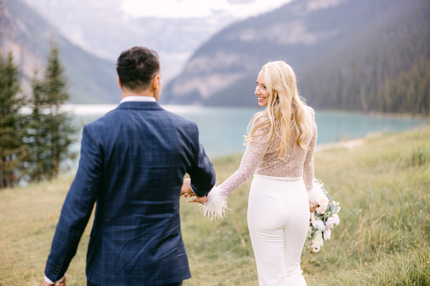 A happy couple walks hand-in-hand by the stunning turquoise waters of Lake Louise, flanked by lush green trees and majestic mountains. The woman, dressed in a sparkly top and white trousers, smiles back at her partner, who wears a stylish suit.