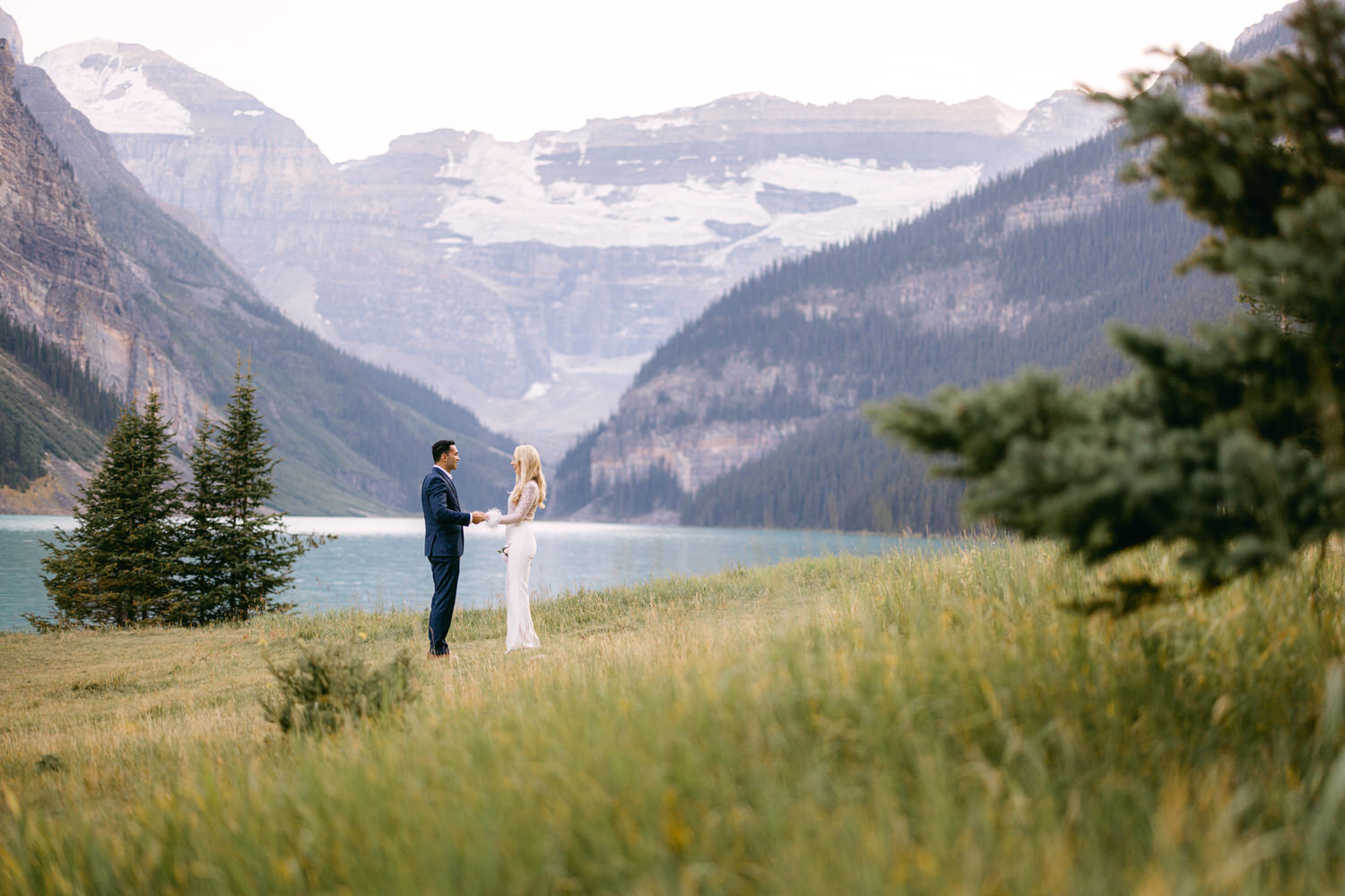 A couple exchanging vows by a serene lake, surrounded by mountains and lush greenery.