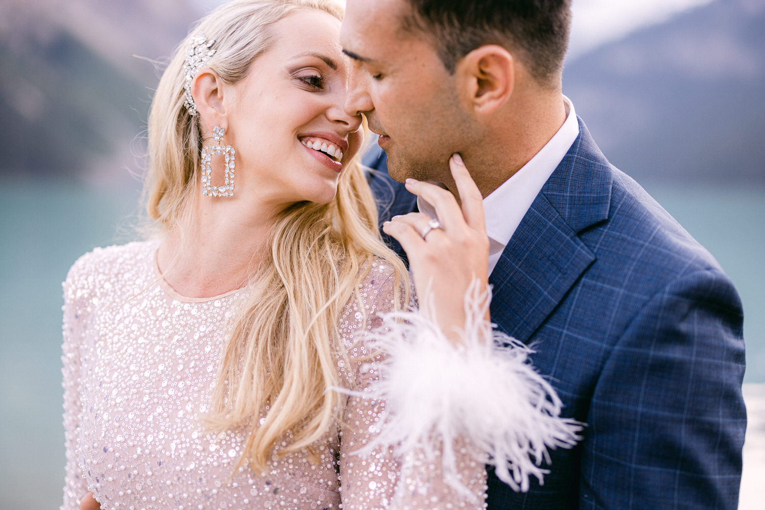 A couple shares a tender moment, with the woman in a sparkling dress adorned with feathers and the man in a stylish suit, set against a serene lakeside backdrop.