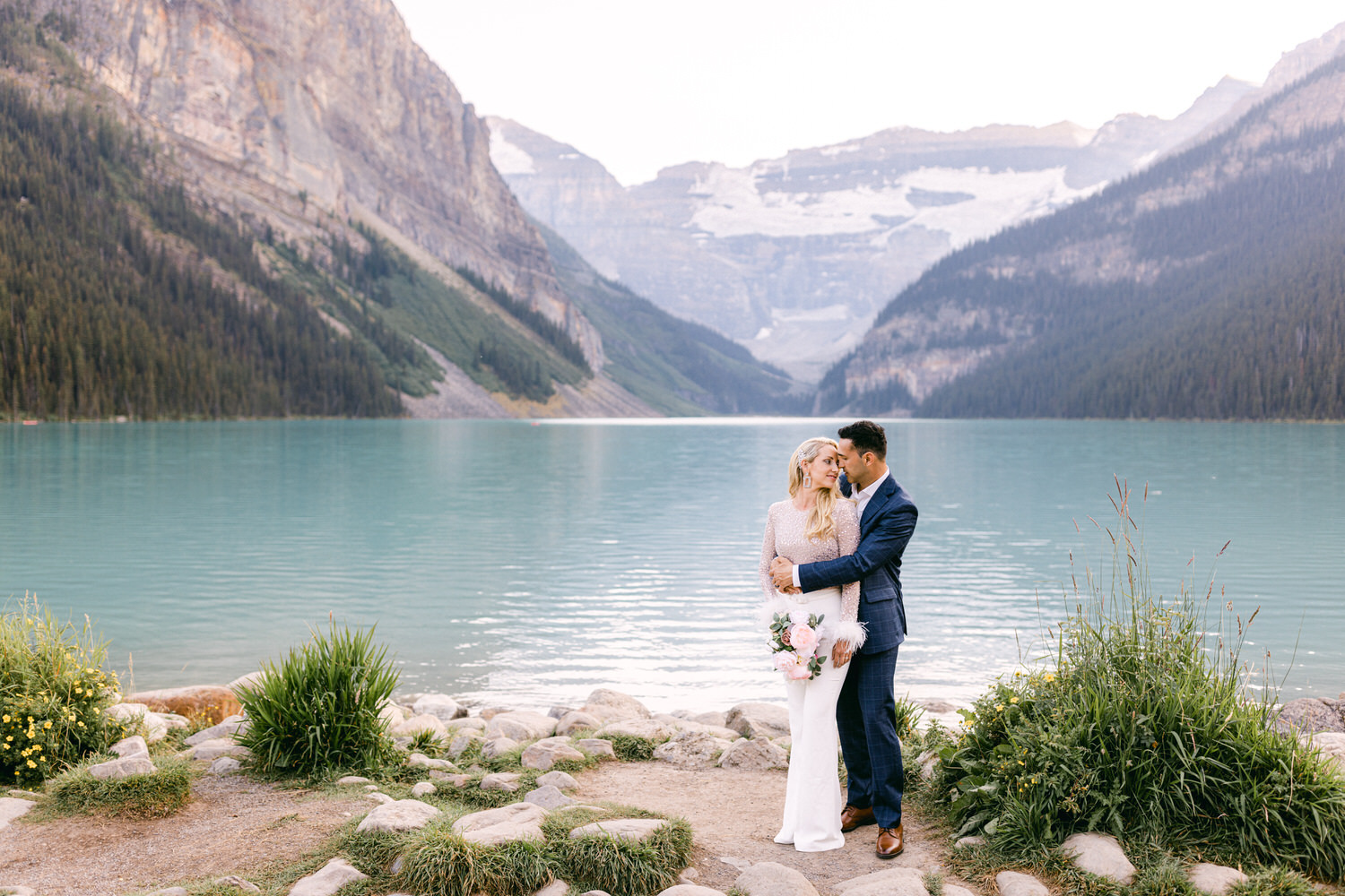 A couple sharing a tender moment by a turquoise lake, surrounded by mountains and lush greenery, with the woman holding a bouquet of flowers.