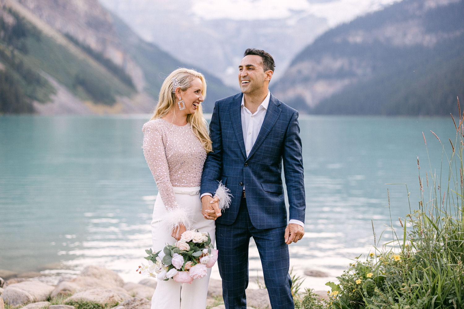 A joyful couple holding hands by a serene lake, surrounded by mountains, with the woman in a sparkling top and the man in a stylish suit, both smiling warmly.