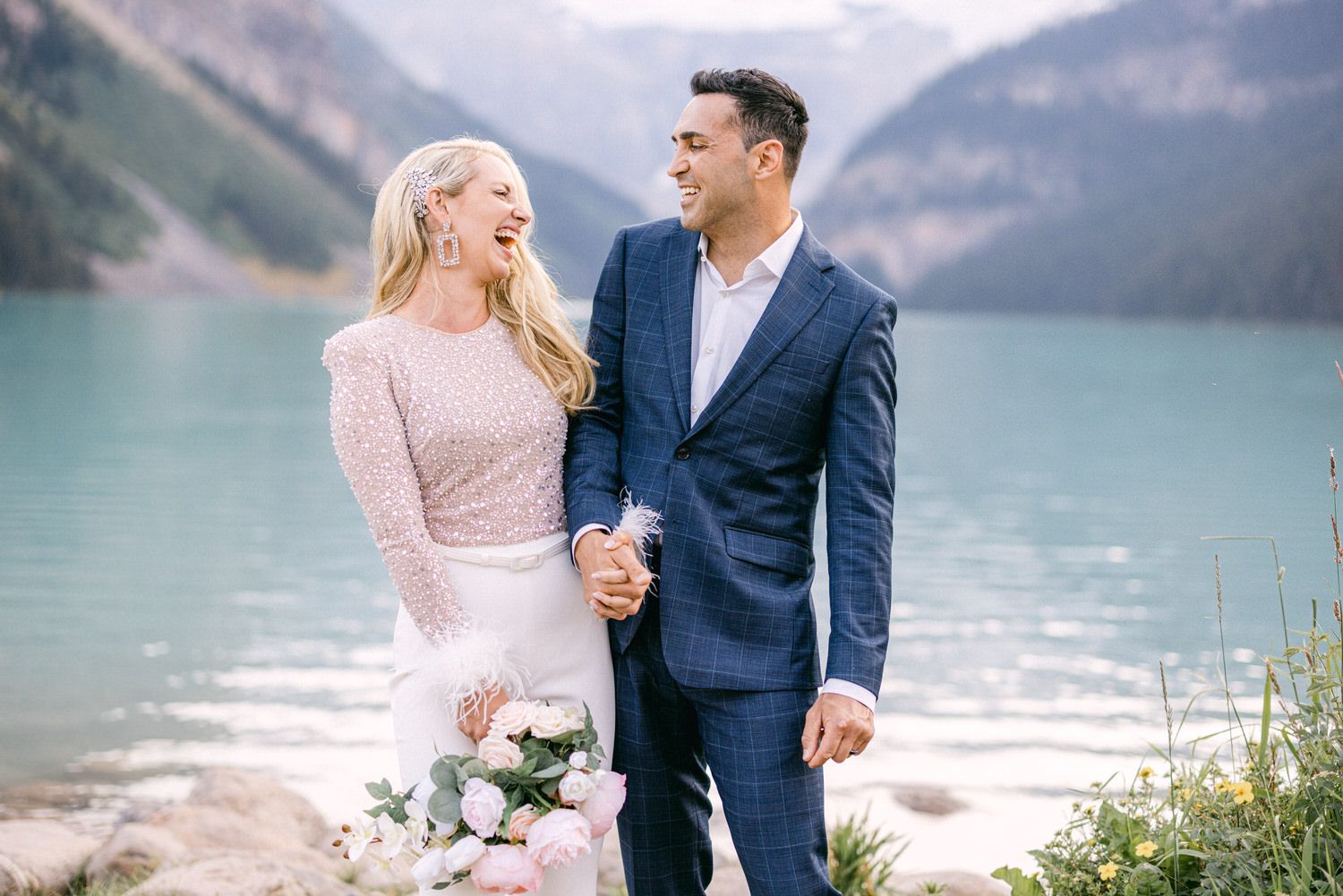 Joyful couple holding hands and laughing together by a serene lake, surrounded by mountains and greenery.