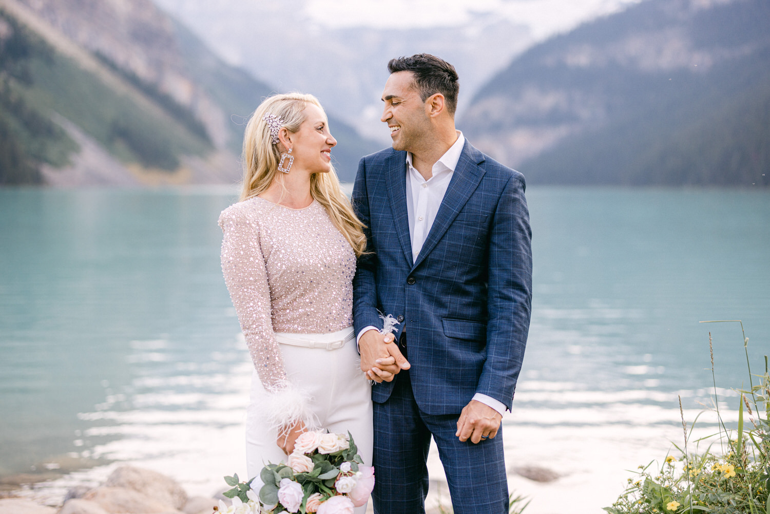 A couple joyfully gazes at each other while standing by a serene lake, adorned in stylish outfits and holding a bouquet of flowers.