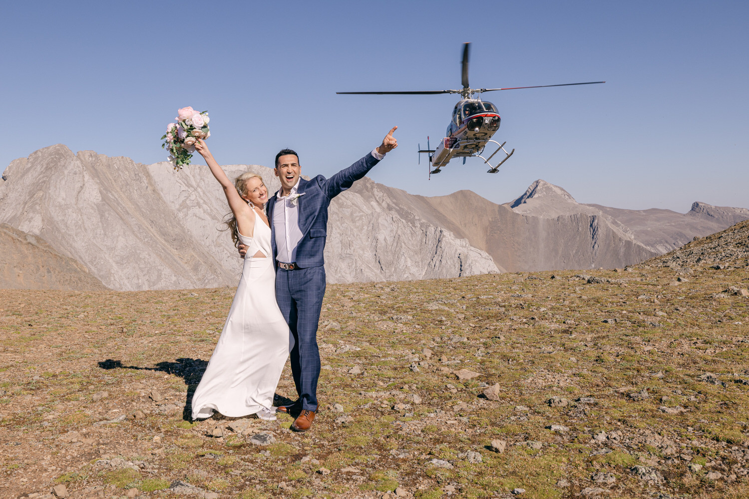 A joyful couple celebrating their wedding on a mountain top with a helicopter flying above, surrounded by stunning natural scenery.