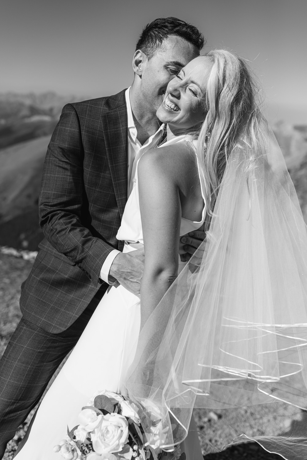 A couple shares a joyful moment on their wedding day, with the groom hugging the bride, both smiling amidst a stunning mountain backdrop. The bride wears a flowing white dress and a veil, while holding a bouquet of flowers.