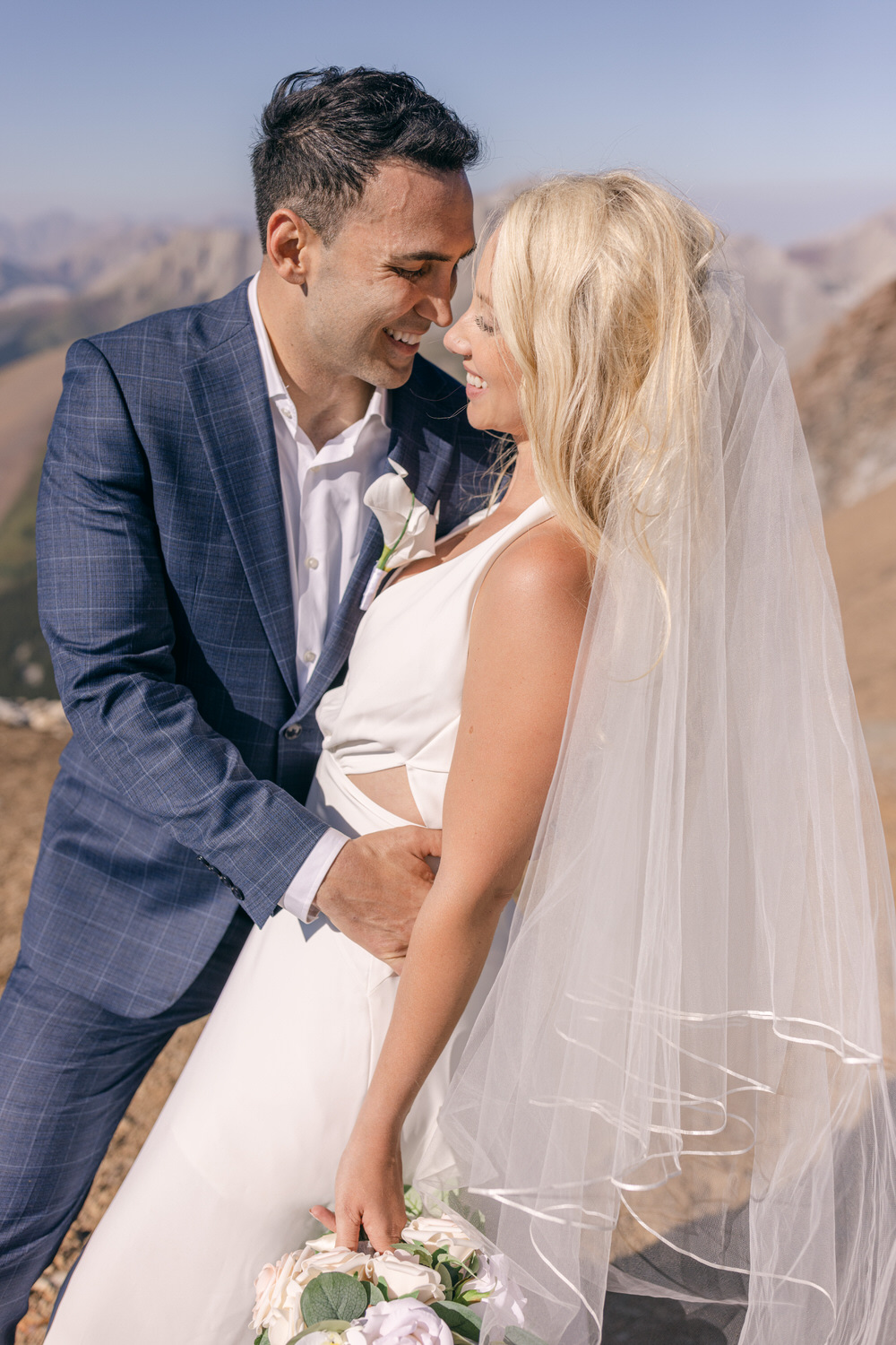 A happy couple embracing on a mountaintop, radiating love and joy, with a bouquet of flowers and a flowing veil.