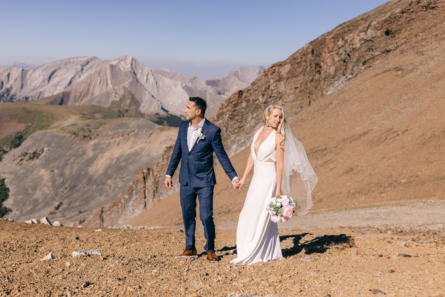 A newlywed couple stands hand in hand on a rocky mountain terrain, with majestic peaks in the background, capturing the essence of their love amidst nature.