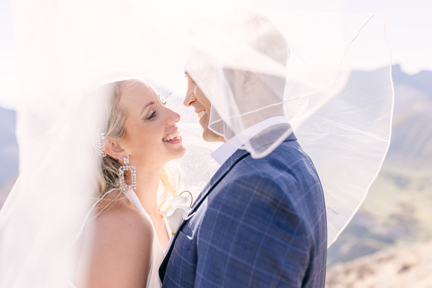 A couple sharing a tender moment under a veil, smiling at each other against a scenic backdrop.