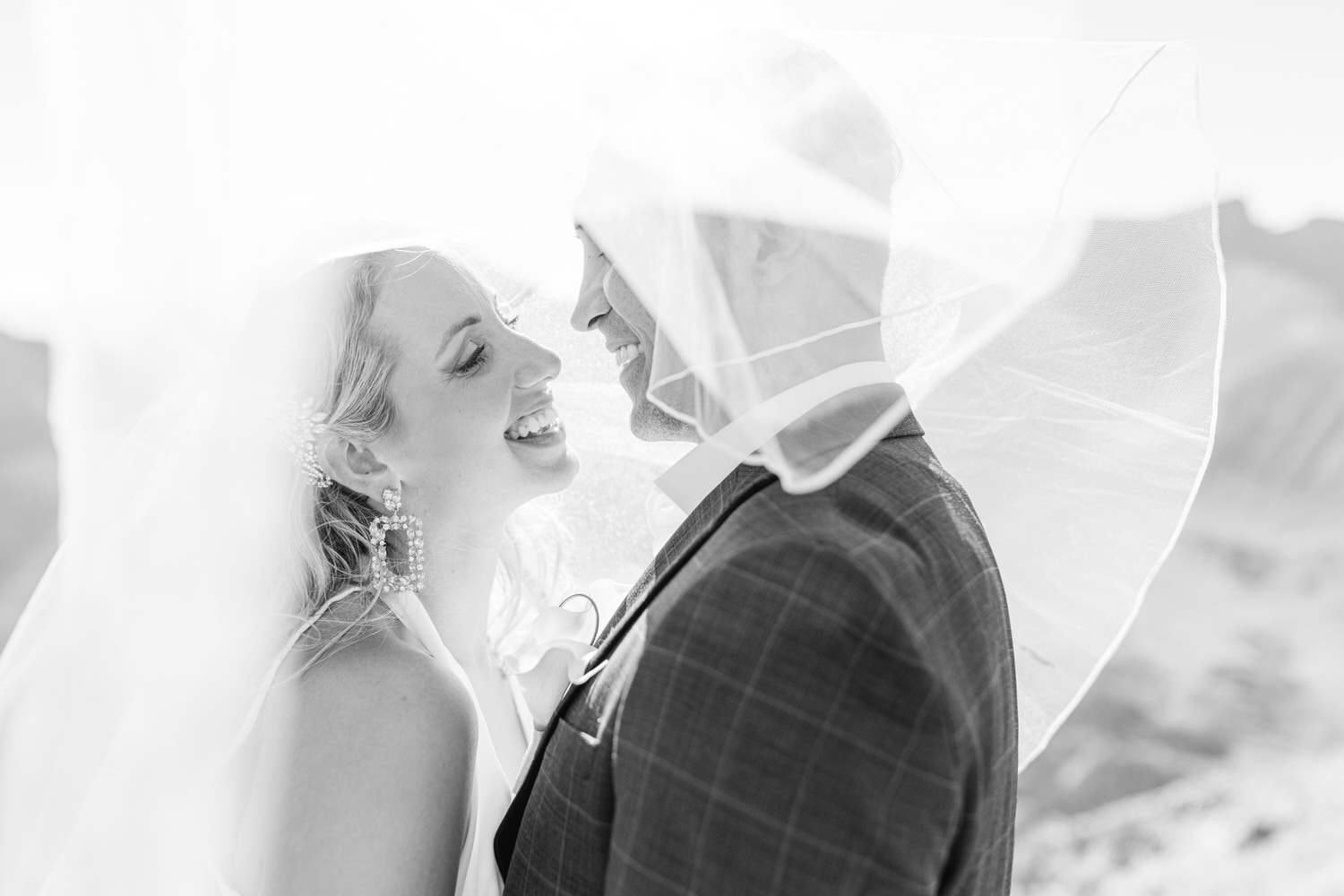 A bride and groom share a joyful smile while their faces are partially covered by a flowing wedding veil, set against a scenic backdrop.