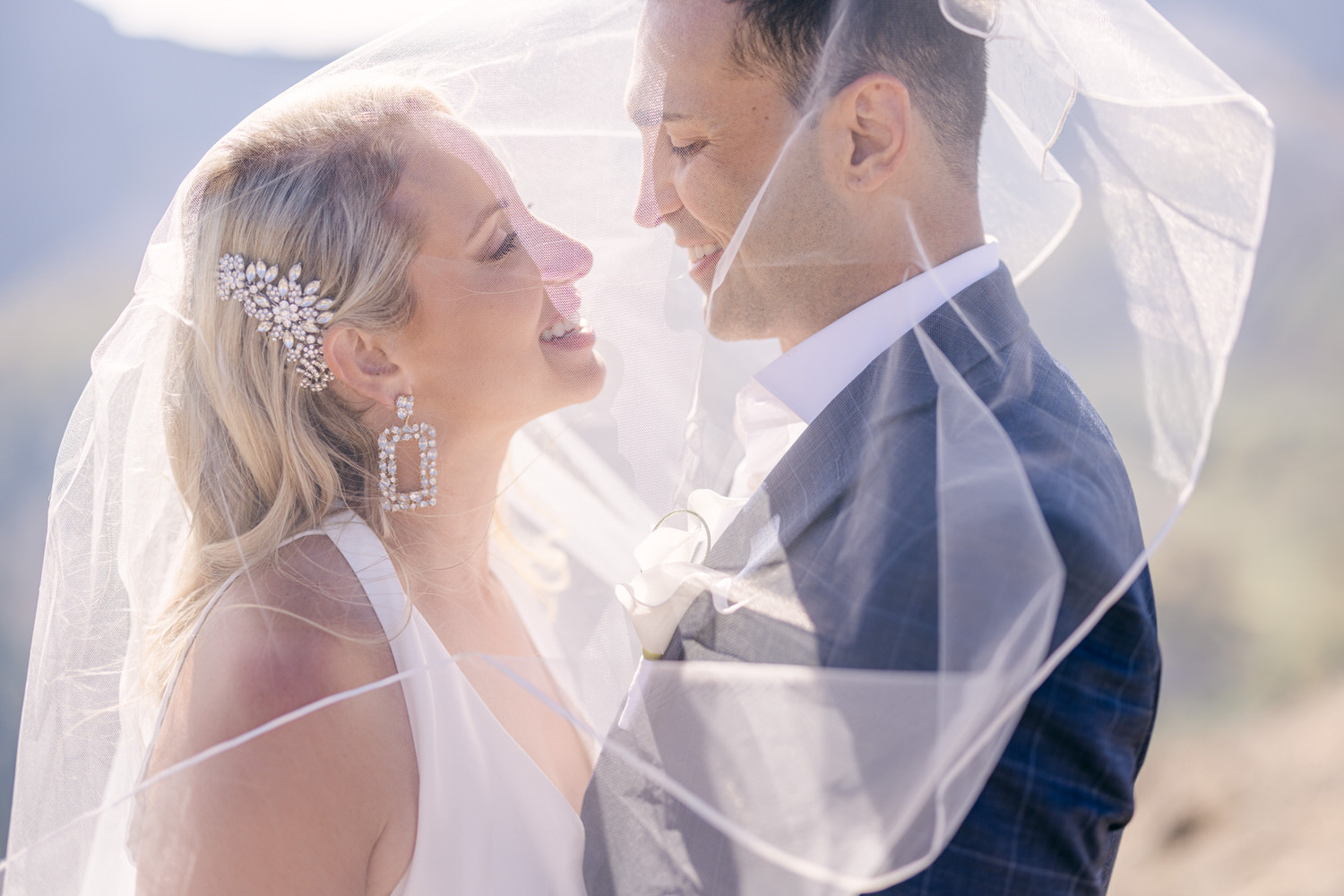 A smiling bride and groom share a joyful moment together, partially obscured by a delicate veil, set against a scenic backdrop.