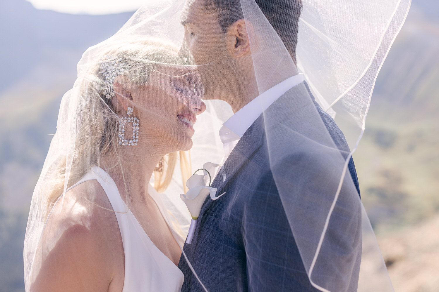 A bride and groom share an intimate moment outdoors, with the bride wearing a shimmering veil and elegant earrings, their foreheads touching as they smile at each other.