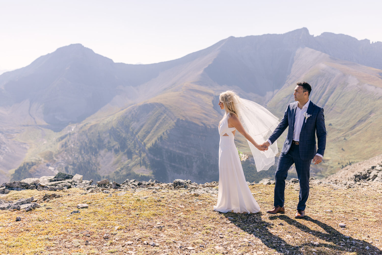 A newlywed couple stands hand in hand on a rocky mountaintop, with breathtaking mountain views in the background.