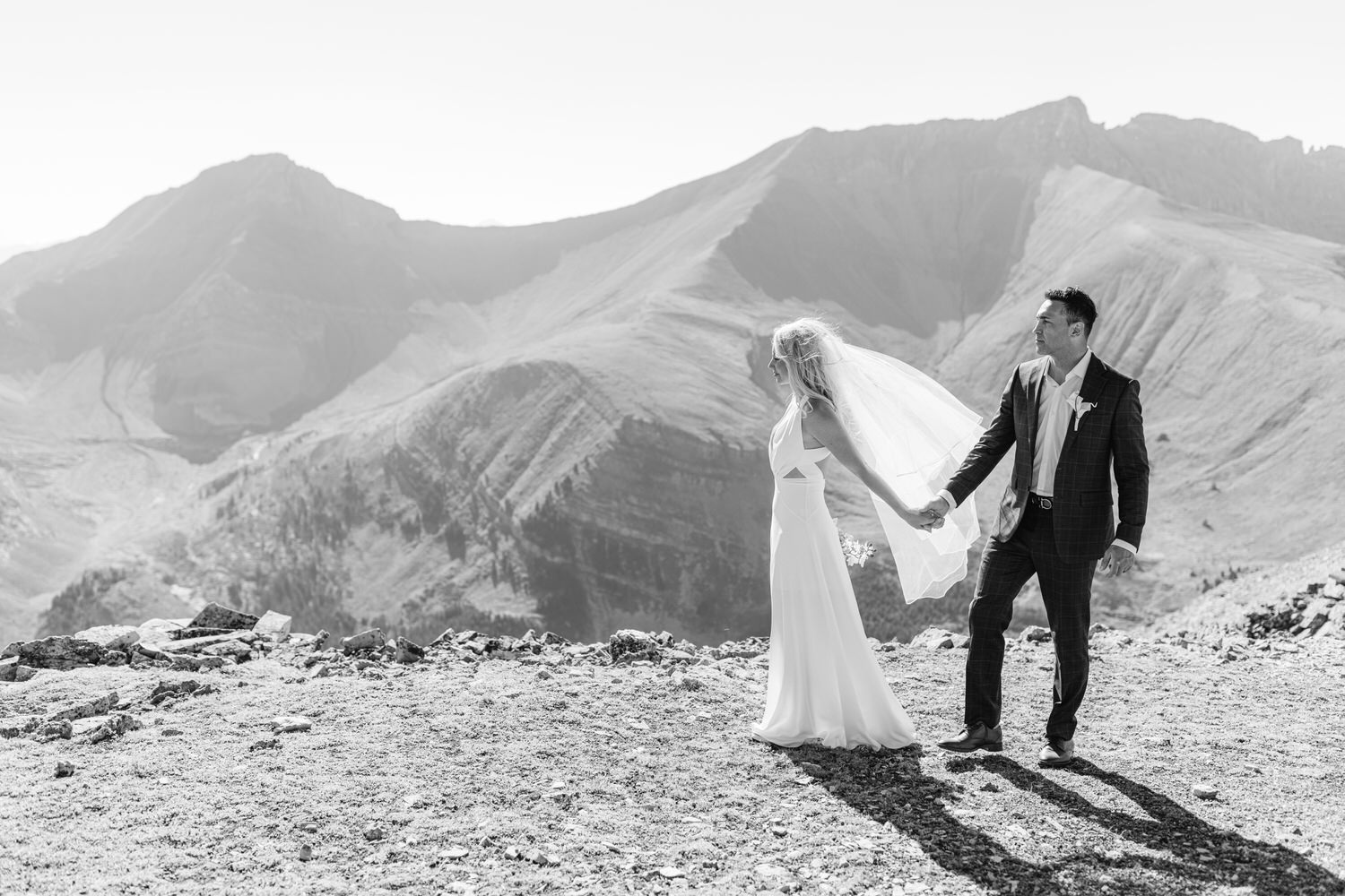 A couple in wedding attire stands hand-in-hand on a rocky outcrop with mountainous scenery in the background, silhouetted against a bright sky.