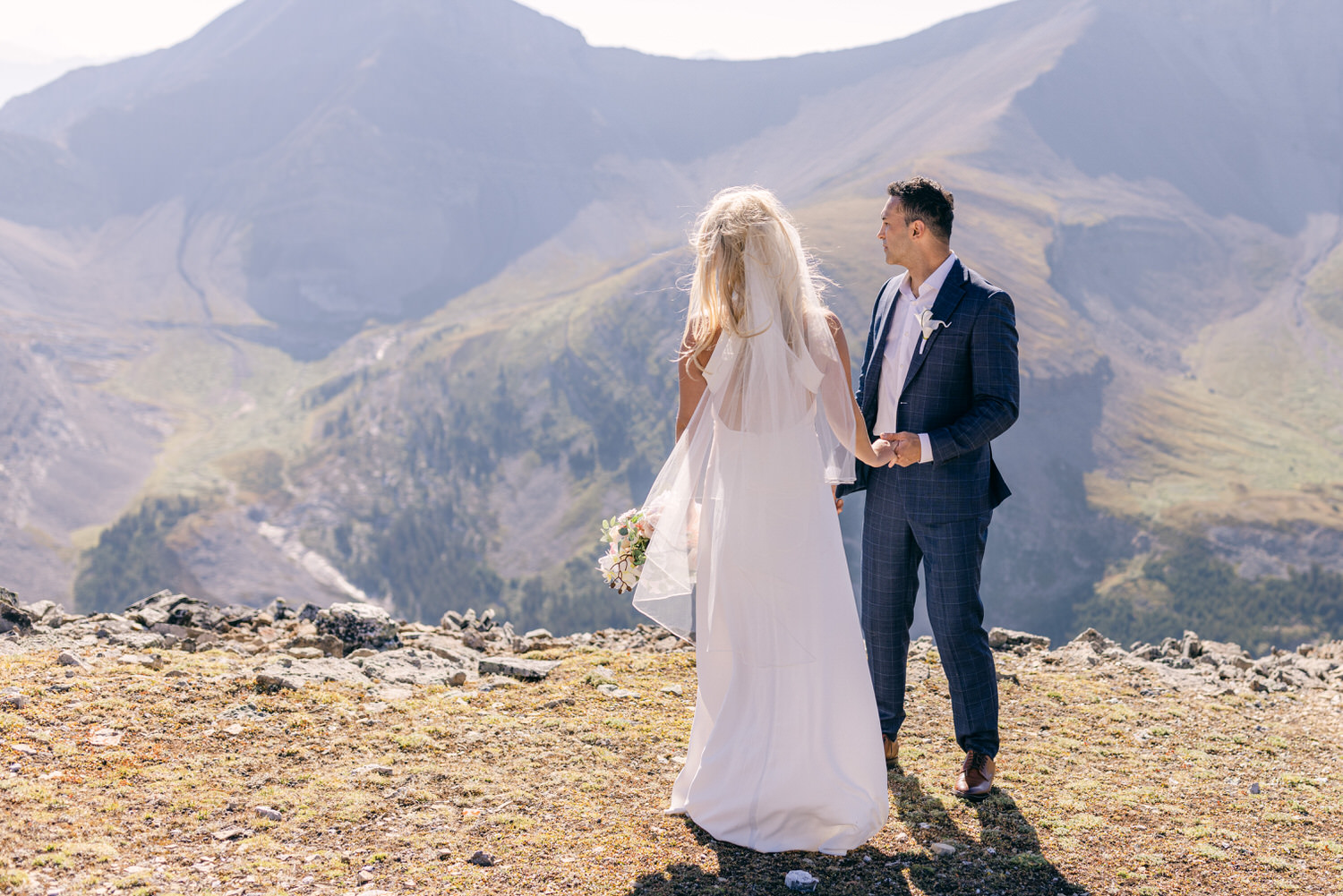 A couple in formal wedding attire stands hand-in-hand on a rocky outcrop with breathtaking mountain views in the background.