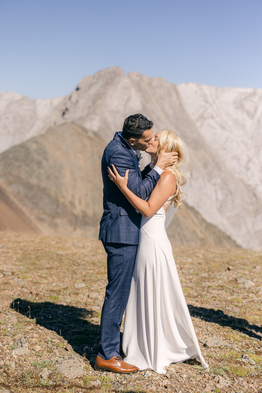 A couple sharing a romantic kiss in wedding attire, set against a stunning mountain backdrop.