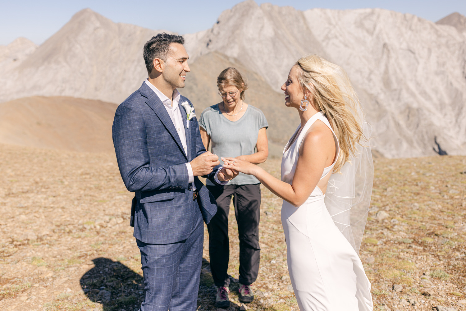 A couple exchanges rings during their wedding ceremony on a scenic mountain top, with a witness present in the background.