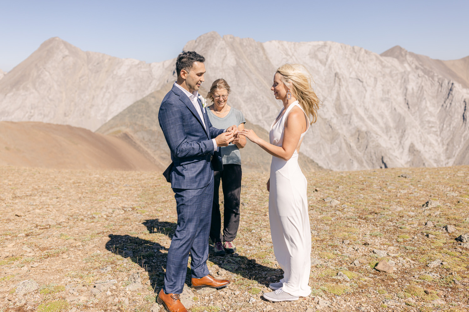 A couple exchanging rings during their outdoor wedding ceremony on a mountain, with a scenic backdrop of rocky peaks and a witness in the background.