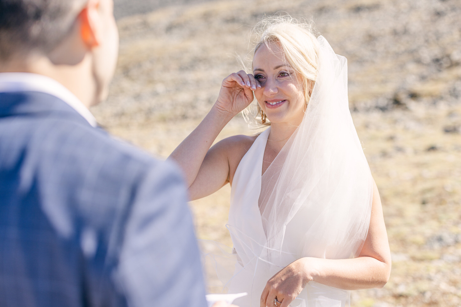 A bride, wearing a white dress and veil, wipes her tears of joy while smiling at her partner during an outdoor ceremony.