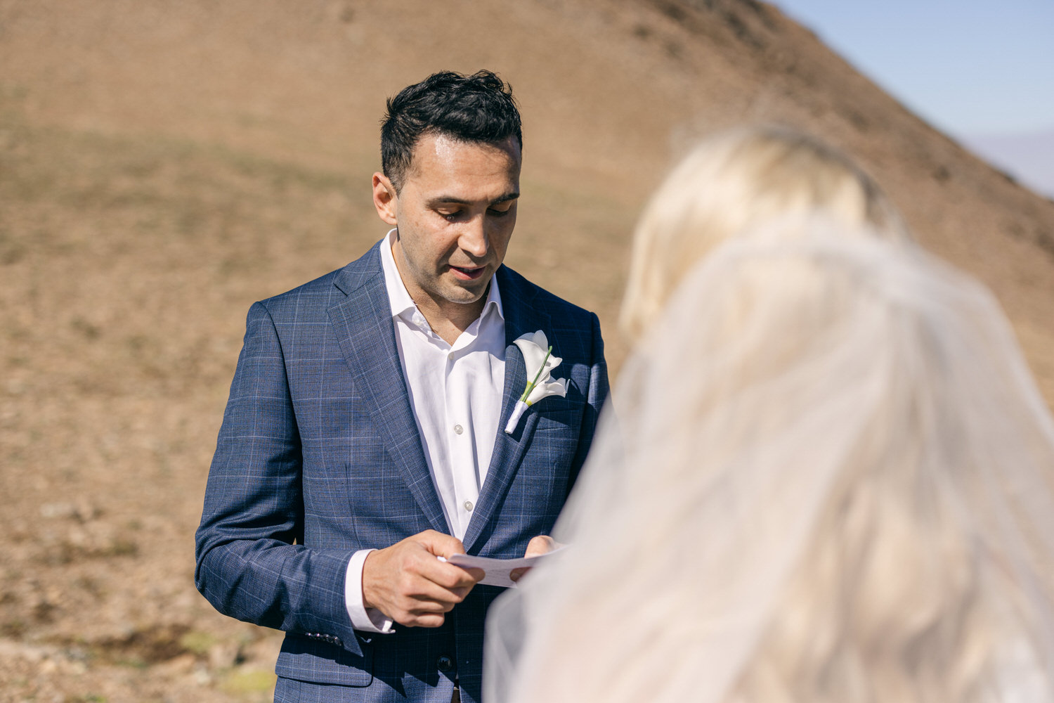 A groom reading vows during an outdoor wedding, with a backdrop of mountains and a bride in the foreground.