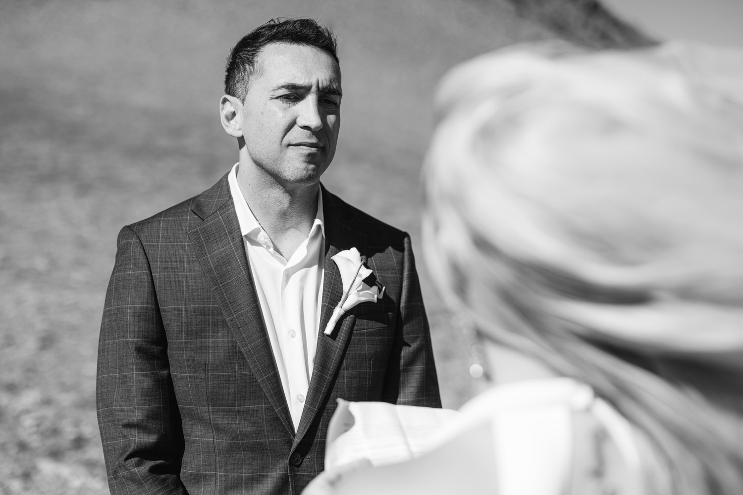 A groom in a suit stands solemnly while a bride, partially visible, holds a wedding document outdoors, with soft wind playing with her hair.