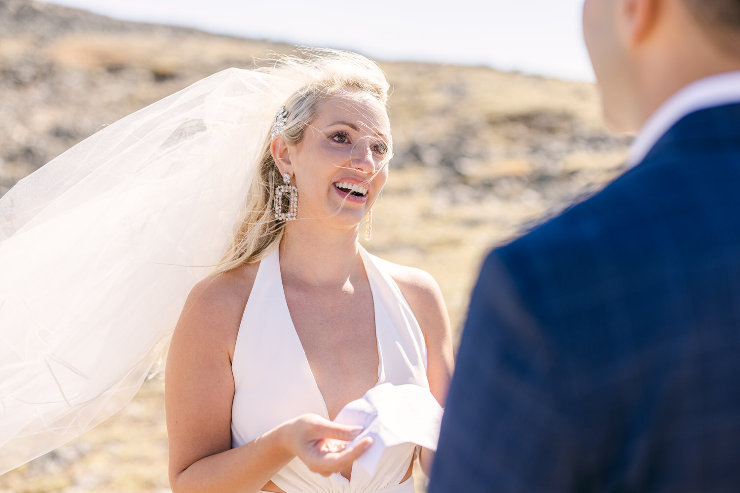 A bride smiles while holding a piece of paper, wearing a white dress and veil, against a scenic outdoor backdrop.