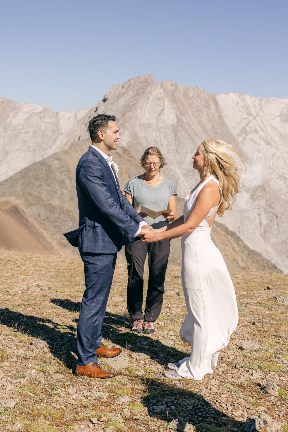 A couple exchanges vows during an outdoor wedding ceremony in a mountainous landscape, with a serene backdrop and an officiant present.