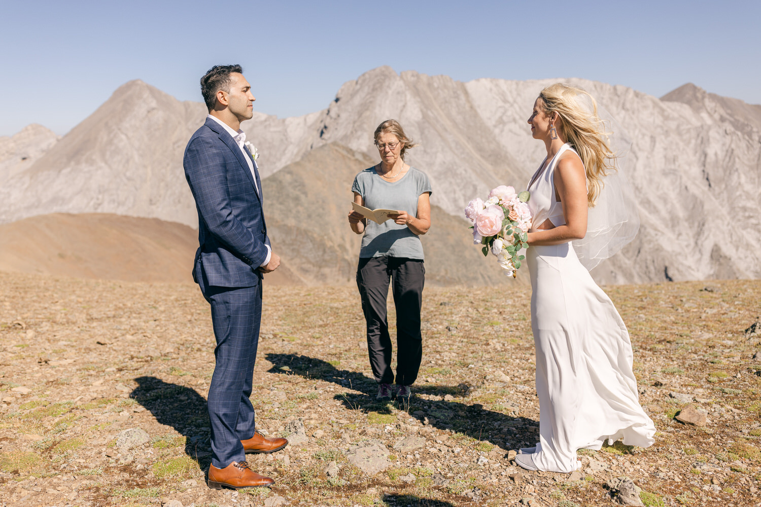 A couple stands hand in hand during their outdoor wedding ceremony in a mountainous landscape, with an officiant reading from a book.