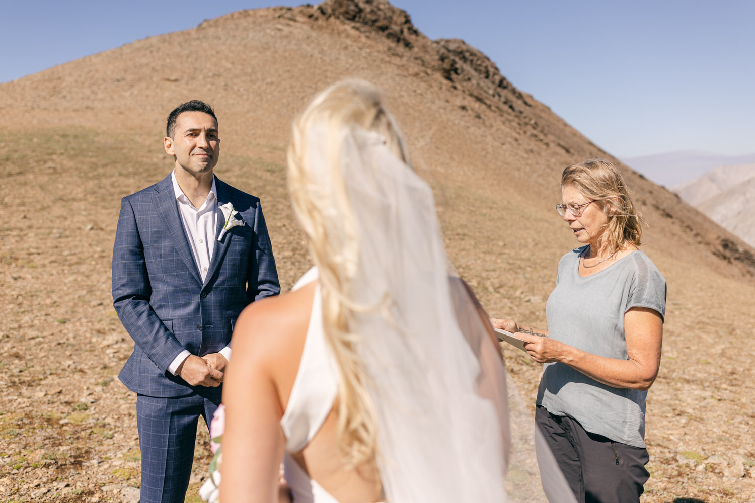 A groom in a suit stands before a woman officiating a wedding on a rocky mountainside, with a bride in a white dress partially visible in the foreground.