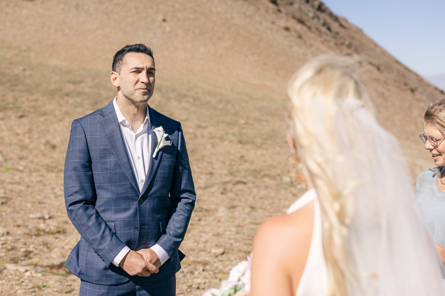 A groom stands thoughtfully during an outdoor wedding ceremony, with a scenic rocky background.