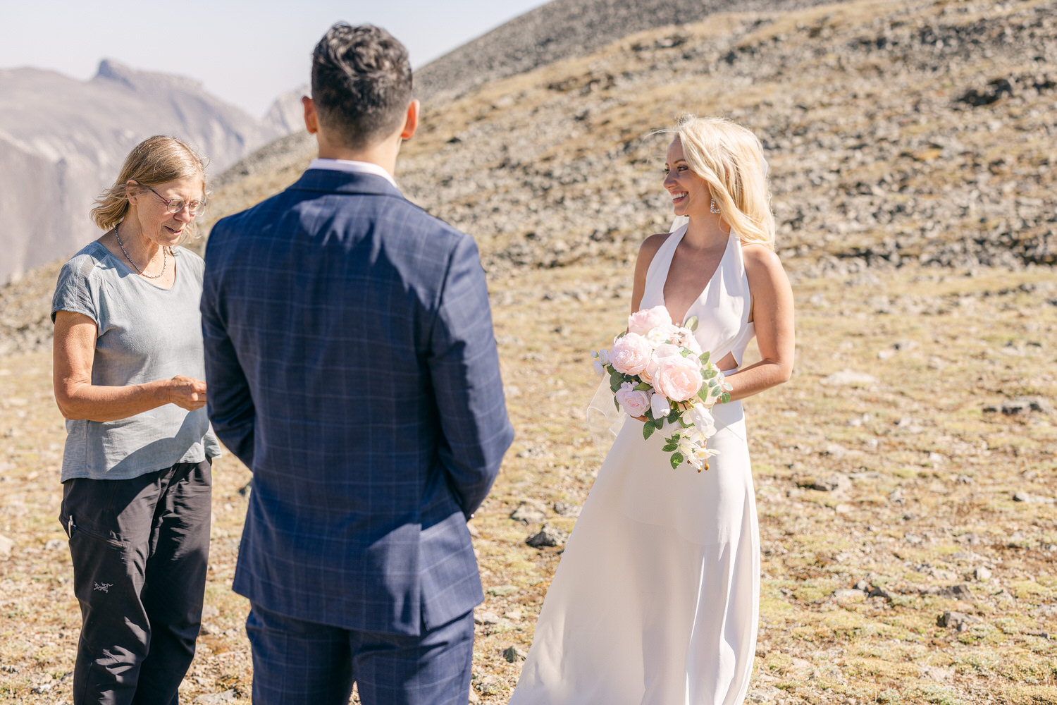 A bride in a flowing white dress smiles joyfully while holding a bouquet of pink roses, as a man in a suit stands in front of her, with a woman overseeing the ceremony in a scenic mountain setting.