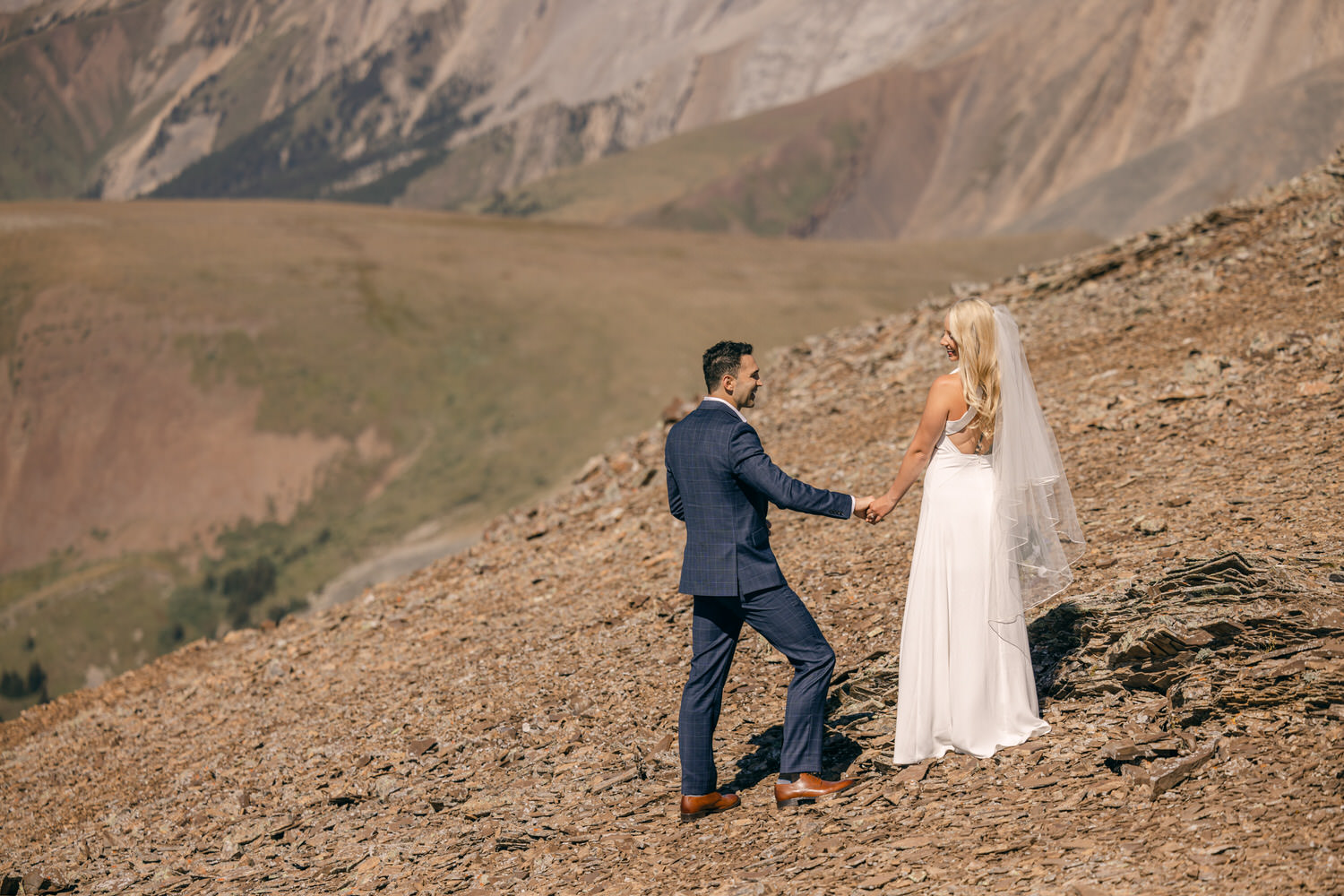 A couple holds hands while standing on rocky terrain with a mountainous backdrop, capturing a serene wedding moment.